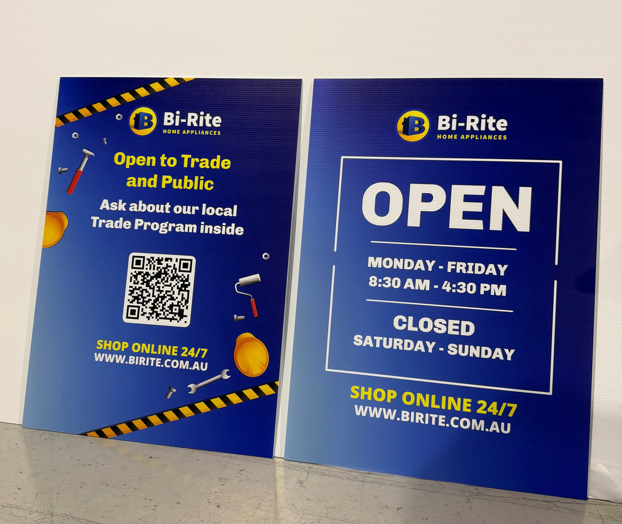 Two Bi-Rite corflute signs on a white wall, one with QR code and text, the other with opening hours.