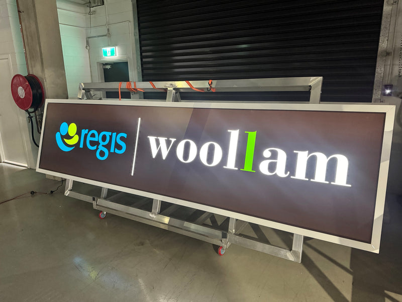 Large Light Box signboard with 'regis woollam' on a stand inside of SS Signs.